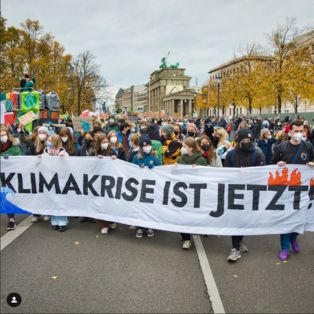 Hinter einem großen Banner mit “Die Klimakrise ist jetzt” laufen viele (vor allem junge) Menschen mit Masken. Sie halten Plakate in die Luft. Im Hintergrund sieht man auch Fridays for Future Flaggen. 