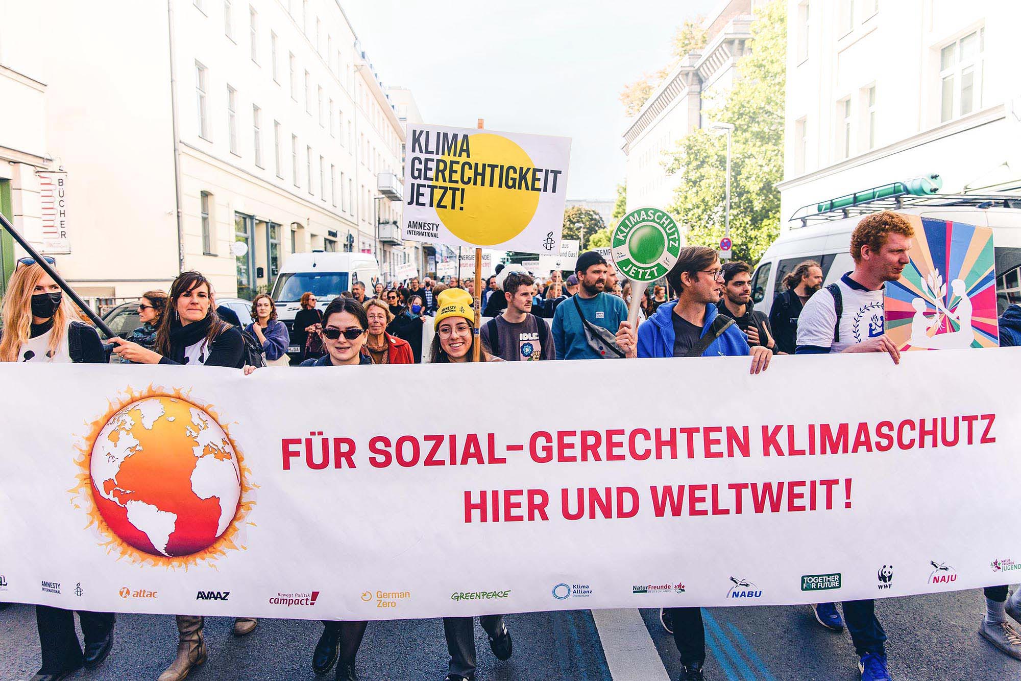 Hinter einem großen Banner mit der Aufschrift: „Für sozial-gerechten Klimaschutz hier und weltweit” stehen Menschen mit bunten Schildern und Plakaten.